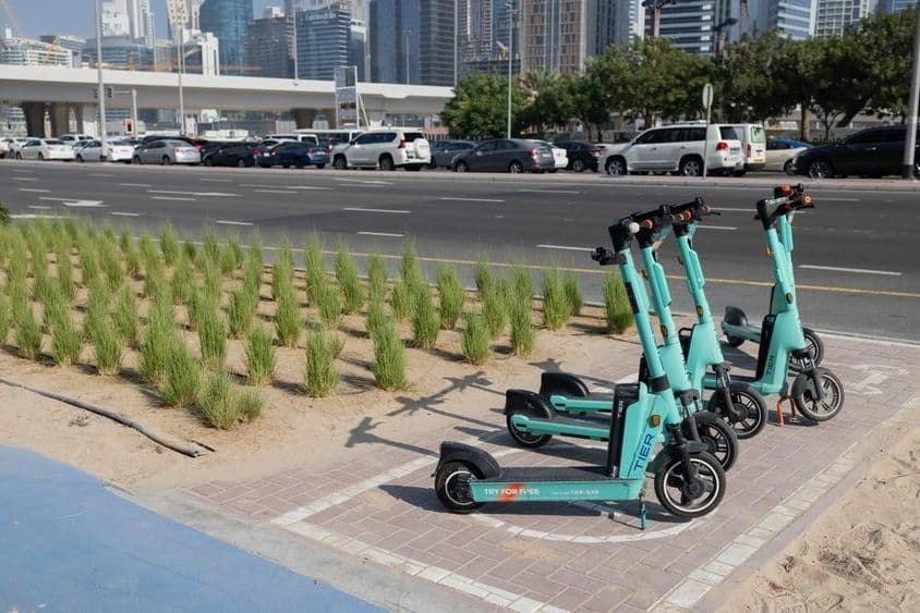 E-scooters estacionados frente a rascacielos de Dubái con plantas verdes en separador de carretera.