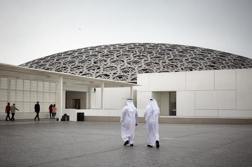Turistas en el museo Louvre Abu Dhabi y dos personas con vestimenta tradicional emiratí blanca.