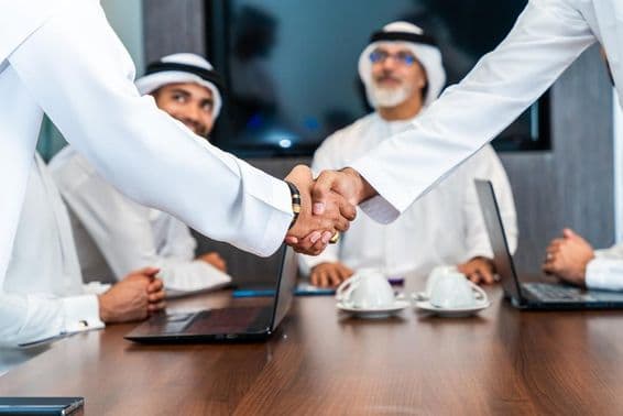 Ejecutivos emiratíes se dan la mano en una mesa de conferencias, con teléfonos, cuadernos y tazas de café sobre la mesa. Dos emiratís sonríen en el fondo.