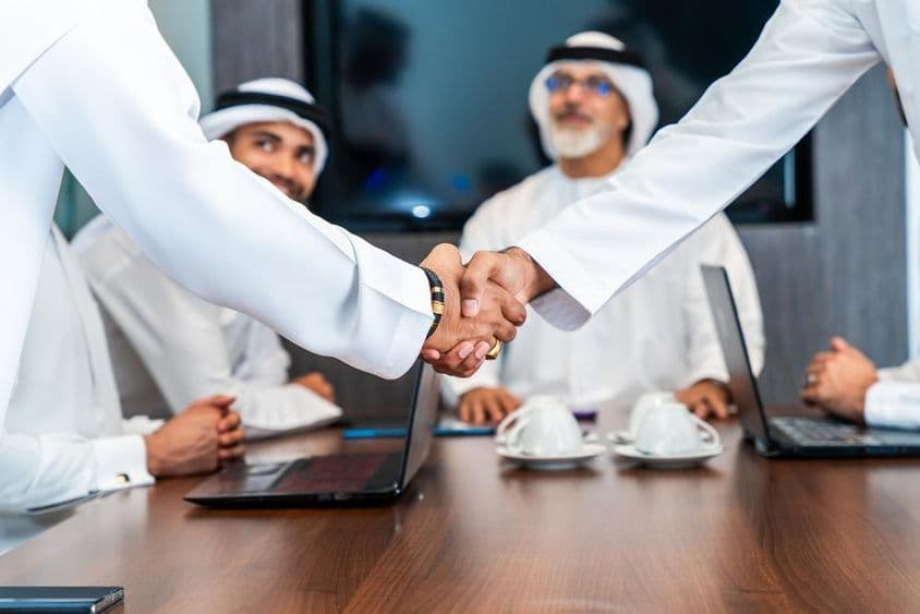 Ejecutivos emiratíes se dan la mano en una mesa de conferencias, con teléfonos, cuadernos y tazas de café sobre la mesa. Dos emiratís sonríen en el fondo.