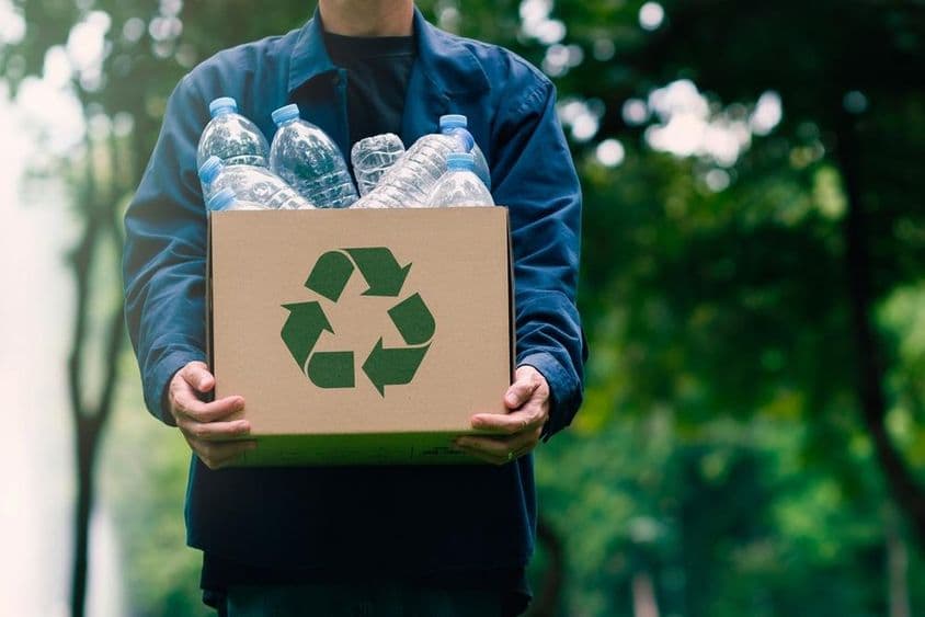 Una persona cargando una caja de cartón con botellas retornables, con árboles verdes desenfocados en el fondo.
