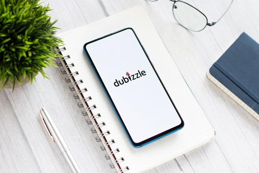 Un teléfono con Dubizzle en una mesa, debajo de un bloc de notas, al lado de gafas y una pequeña planta verde.