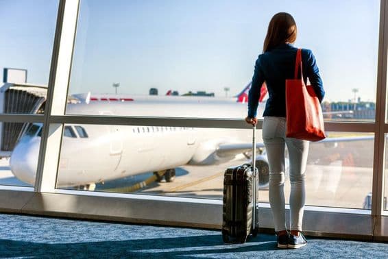 Una mujer de pie junto a la ventana con un avión visible, sosteniendo una maleta con ruedas con su mano izquierda y un bolso de mano en la derecha.