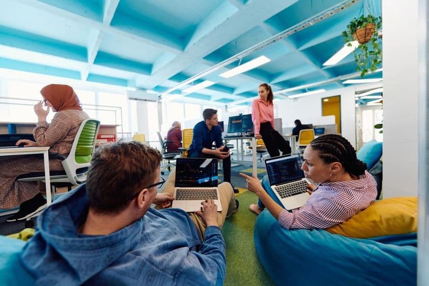 Personas trabajando en una oficina, dos individuos sentados en pufs con portátiles sobre sus regazos.
