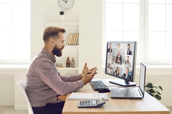 Una persona en un entorno de oficina con un cuaderno y una calculadora sobre el escritorio, realizando una videoconferencia en un monitor con cámara web.