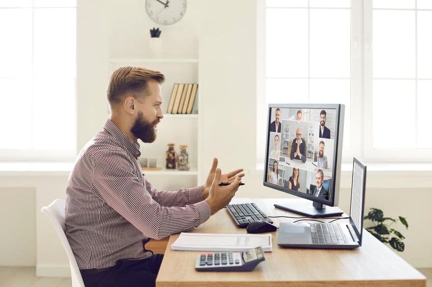 Una persona en un entorno de oficina con un cuaderno y una calculadora sobre el escritorio, realizando una videoconferencia en un monitor con cámara web.