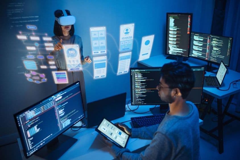 Un desarrollador de aplicaciones sentado frente a una computadora, sosteniendo una tableta, con dos monitores mostrando código de computadora.