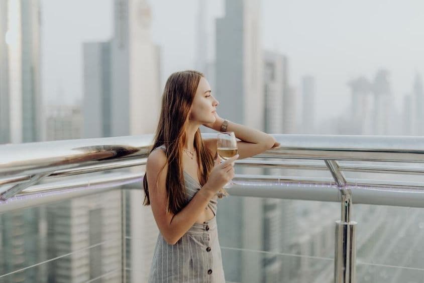 Joven en una terraza con una copa de vino, rascacielos de Dubai borrosos de fondo.