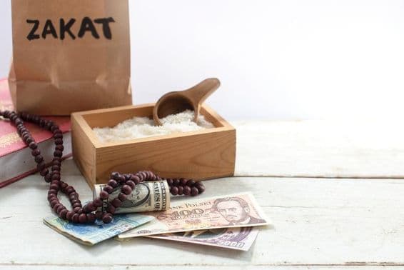 Bolsa etiquetada 'Zakat' frente a una caja con arroz, perlas, dólares y otras monedas.