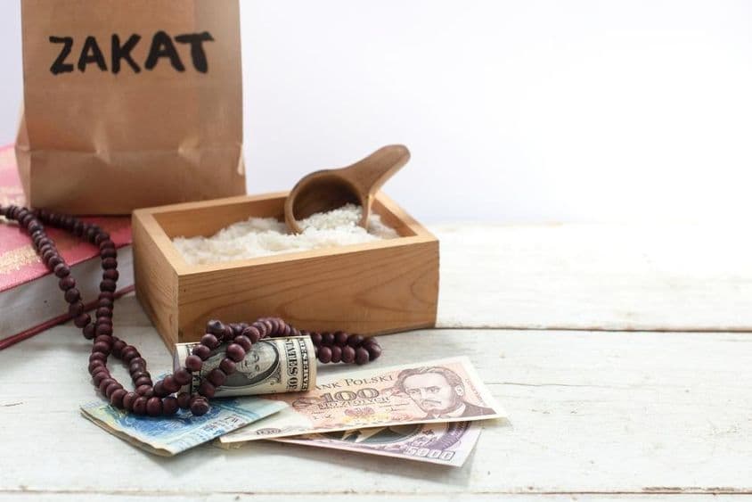 Bolsa etiquetada 'Zakat' frente a una caja con arroz, perlas, dólares y otras monedas.