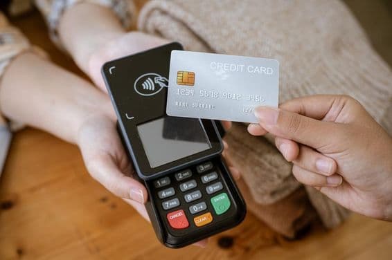 Una tarjeta bancaria sostenida por una mano, con un lector de tarjetas al fondo.