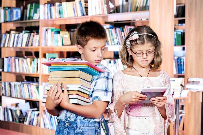 Dos niños en la biblioteca, uno leyendo un libro electrónico, el otro cargando muchos libros en sus manos.