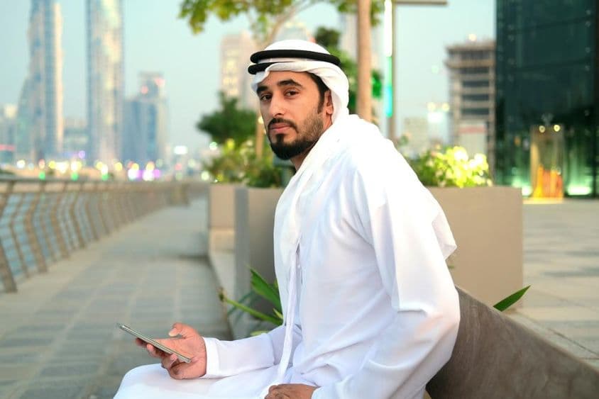 Ciudadano emiratí sentado en un banco, sosteniendo un teléfono, rascacielos al fondo.
