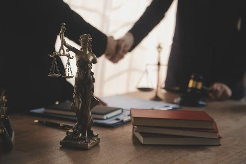 Un abogado se muestra negociando con un cliente, con una estatua de Justicia visible sobre la mesa.
