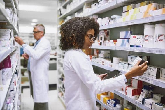 Un estante de farmacia siendo abastecido por una mujer y un hombre revisando medicamentos.