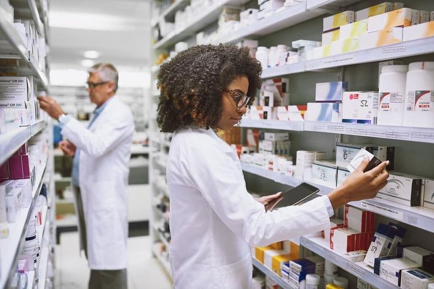 Un estante de farmacia siendo abastecido por una mujer y un hombre revisando medicamentos.