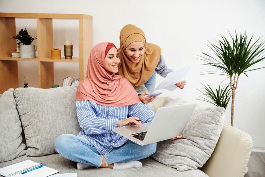 Dos mujeres emiratíes revisando un contrato y un cuaderno, una sosteniendo un teléfono, sentadas en un sofá.