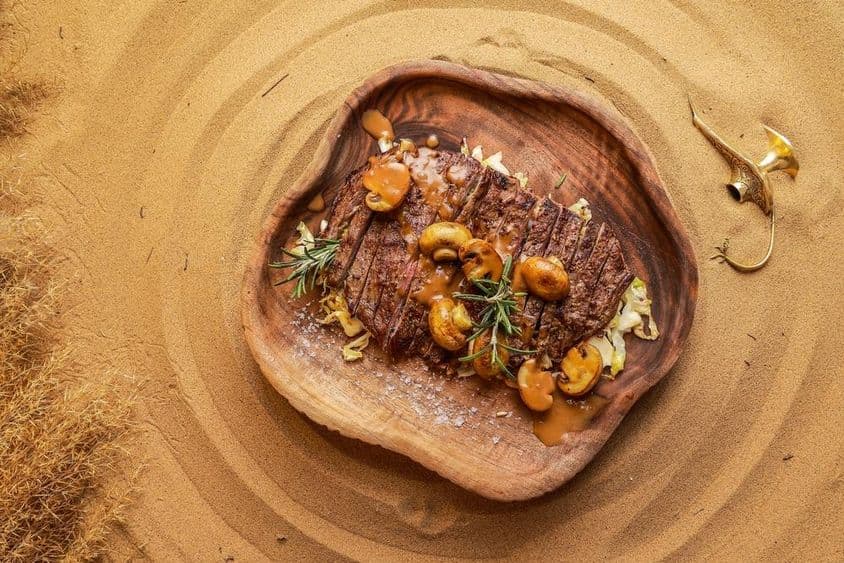 Bistec servido con champiñones en un plato de madera en la arena del desierto, con una linterna apagada al lado.