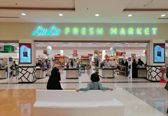 Entrada a una tienda LuLu en un centro comercial, con dos personas sentadas, una mujer y un niño, vistos desde atrás.