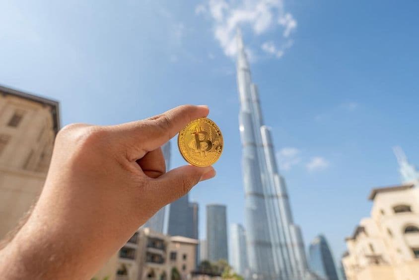 Persona sosteniendo una moneda de oro de Bitcoin, mirando al cielo, con el Burj Khalifa de fondo.
