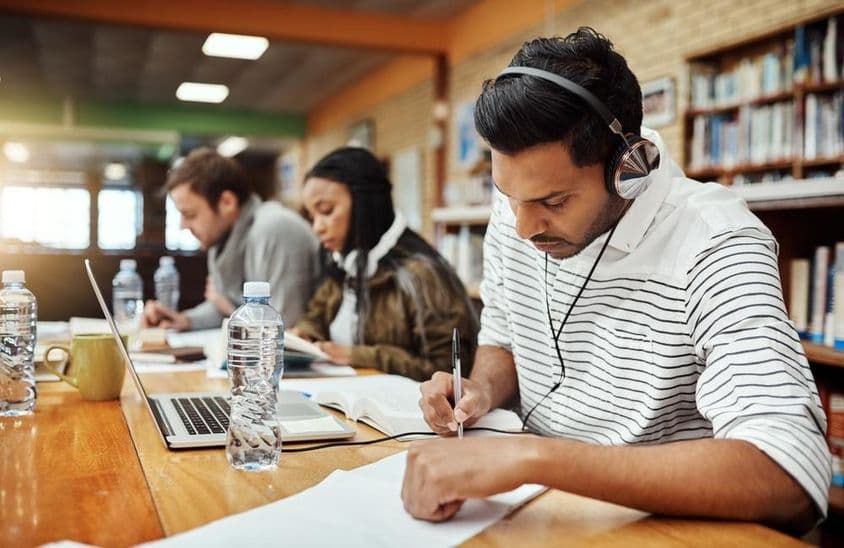 Estudiantes universitarios escribiendo en papel con libro y cuaderno enfrente, uno con auriculares puestos.