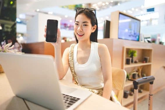 Chica de la Generación Z trabajando, sosteniendo un teléfono, con un cuaderno frente a ella y una asa de maleta a su lado.