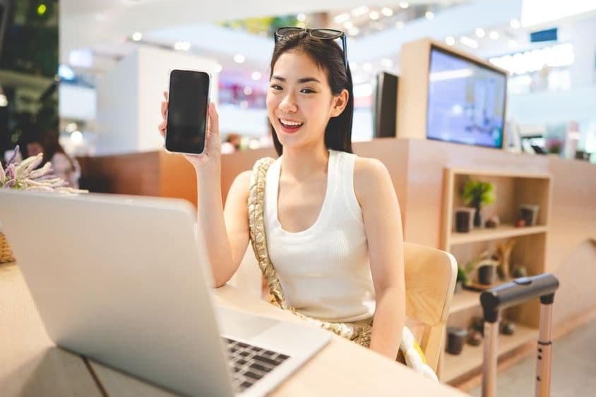 Chica de la Generación Z trabajando, sosteniendo un teléfono, con un cuaderno frente a ella y una asa de maleta a su lado.