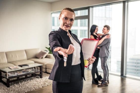 Una corredora de bienes raíces saludando, una pareja abrazándose al fondo.