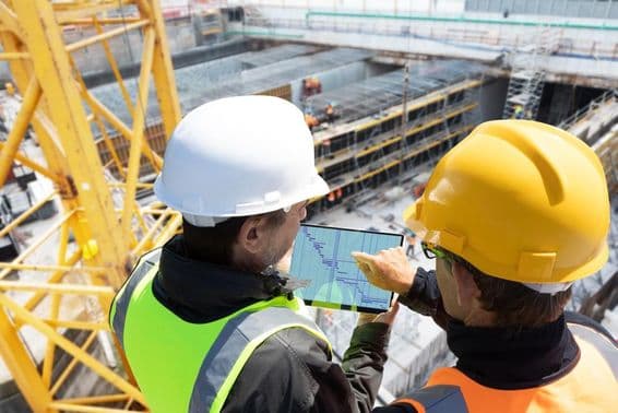 Dos personas con cascos en una grúa en un sitio de construcción, revisando planos en una tableta, con el sitio de construcción frente a ellos.