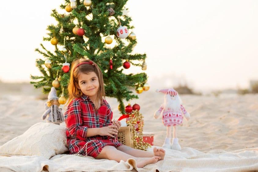 Niña pequeña sonriente sentada en la playa bajo un árbol de Navidad.