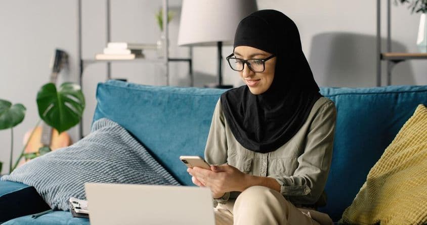 Chica árabe en hijab trabajando en casa usando smartphone y laptop mientras está sentada en el sofá.