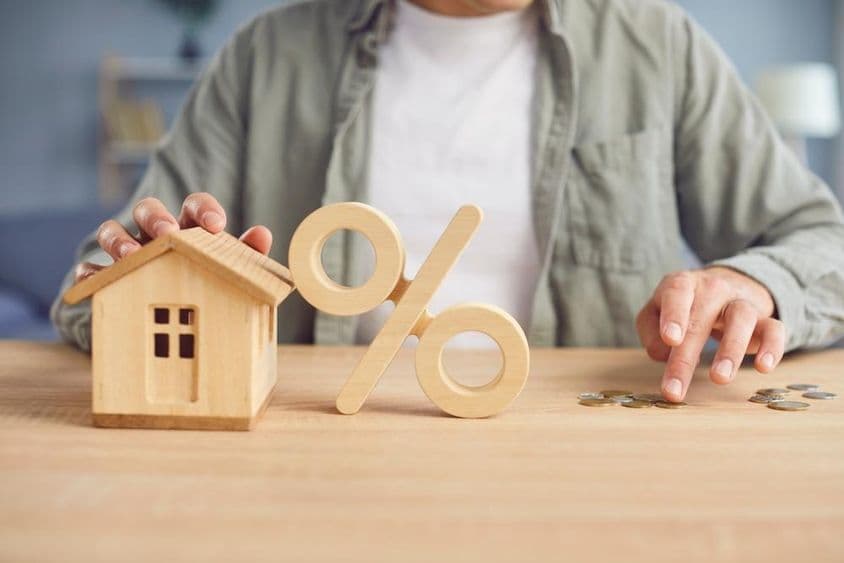 Símbolo porcentual de madera y modelo de casa sobre una mesa con un joven contando monedas en el fondo.