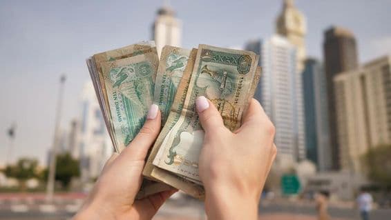 Una niña sosteniendo moneda de Emiratos Árabes Unidos con rascacielos de Dubái al fondo.