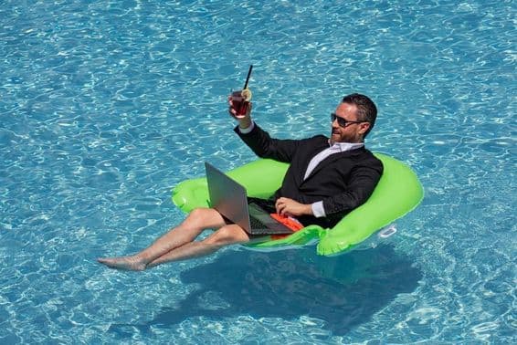 Hombre de negocios exitoso flotando en una piscina con traje, bebiendo un cóctel de verano y usando una laptop en la piscina.