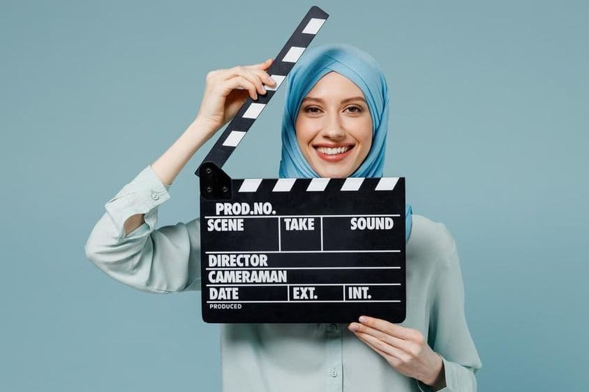 Mujer árabe musulmana joven en abaya y hiyab sosteniendo una claqueta de cine clásica negra.