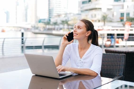 Joven mujer freelance trabajando en una laptop, sentada en una cafetería al aire libre sosteniendo un teléfono.