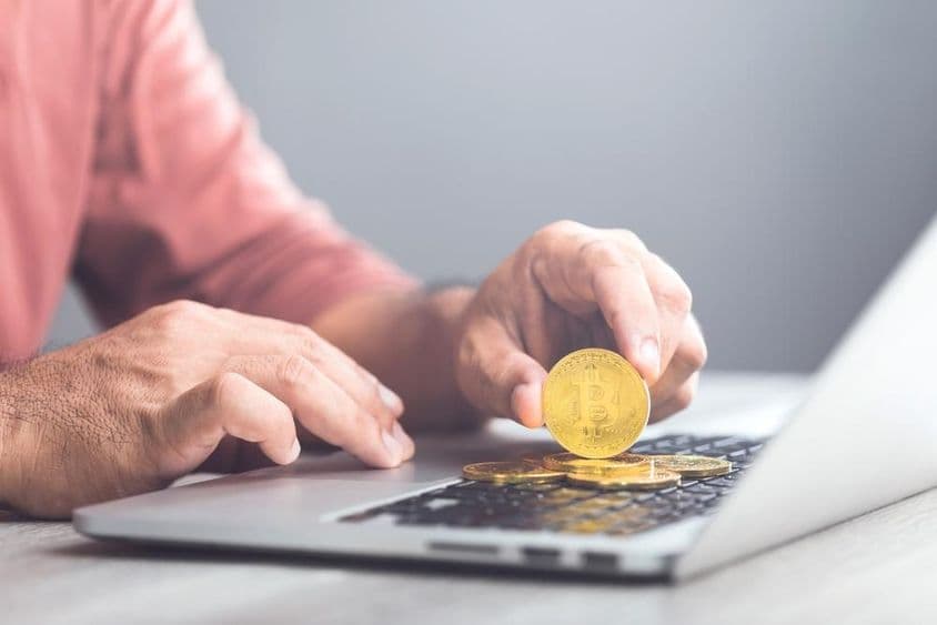 Hombre sosteniendo un Bitcoin color dorado mientras usa una laptop.