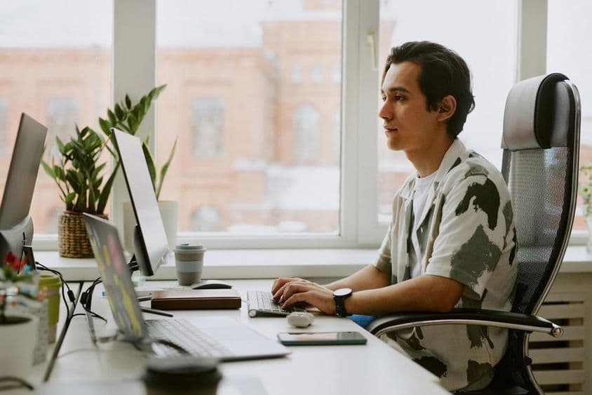 Joven programador sentado en un escritorio, codificando en una computadora en una oficina luminosa.