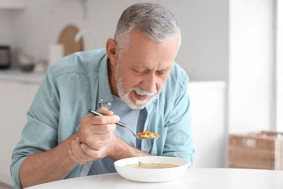 Hombre mayor con Parkinson comiendo en una mesa de cocina.