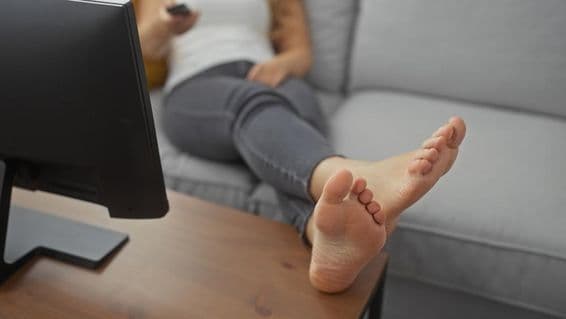 Una joven descansando descalza en un cómodo sofá de la sala, sosteniendo un control remoto.