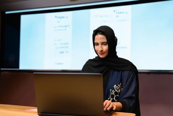 Maestra con Abaya Hijab frente a pantalla de proyector en un aula moderna con pantalla digital.