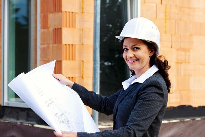 Arquitecta con plano frente a una casa en construcción, usando casco de seguridad.