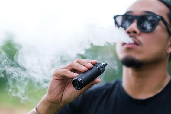 Hombre asiático fumando con un cigarrillo electrónico de vapeo, exhalando humo mientras disfruta al aire libre en verano.