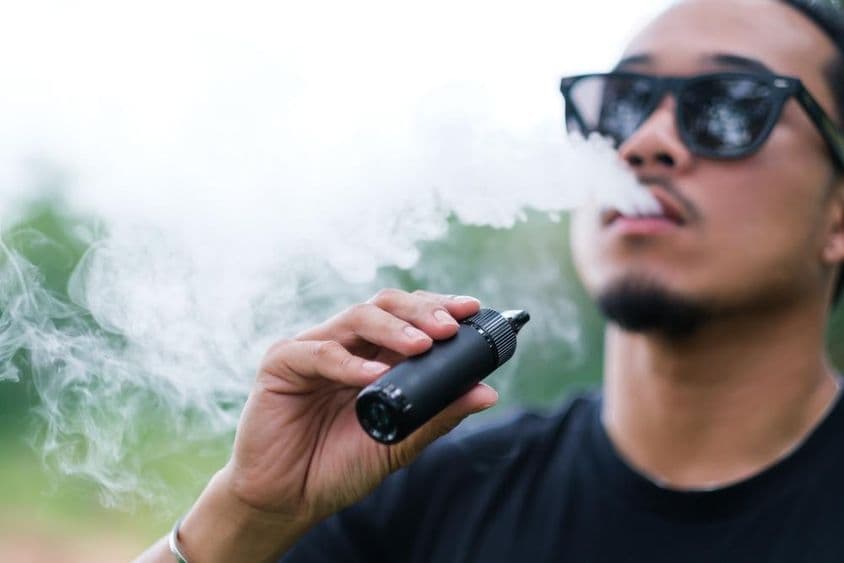Hombre asiático fumando con un cigarrillo electrónico de vapeo, exhalando humo mientras disfruta al aire libre en verano.