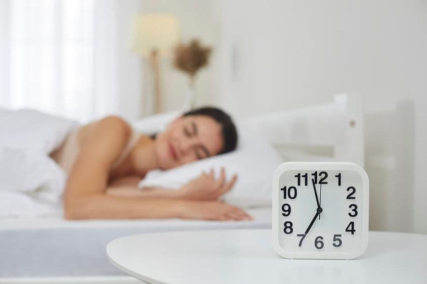 Reloj despertador en una mesa de noche en un dormitorio, con una joven durmiendo en el fondo.