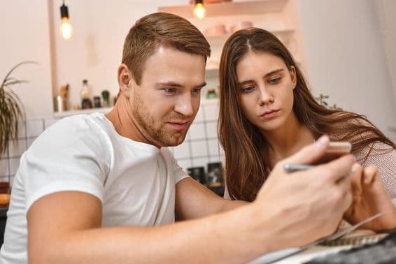 Pareja joven disfrutando un café en la cocina mientras leen una notificación bancaria en línea en un teléfono móvil.