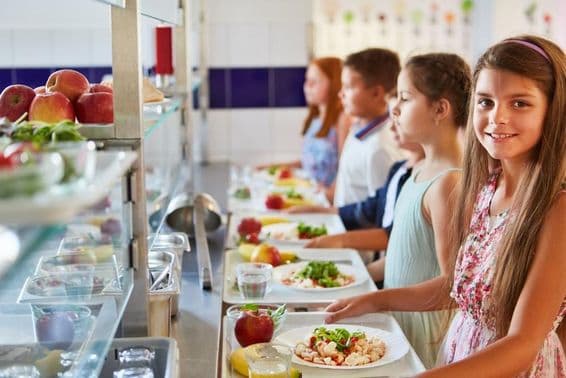 Niña sonriente con bandeja de comida, en fila con amigos durante el recreo en la cafetería escolar.