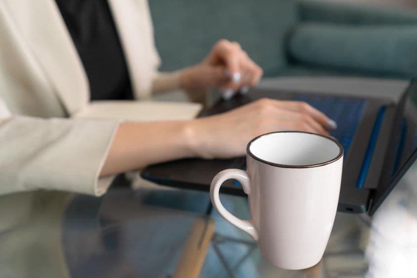 Una taza sobre una mesa de cristal cerca de una laptop, con una joven escribiendo.