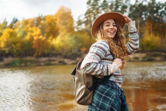 Viajera con mochila y gorra disfrutando de una caminata otoñal junto a un río.