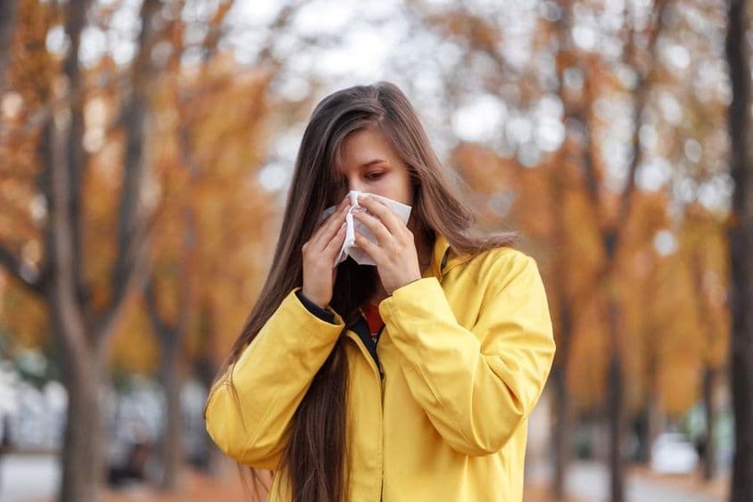 Mujer joven de pie en un parque con un abrigo amarillo, sosteniendo un pañuelo en la nariz mientras estornuda.